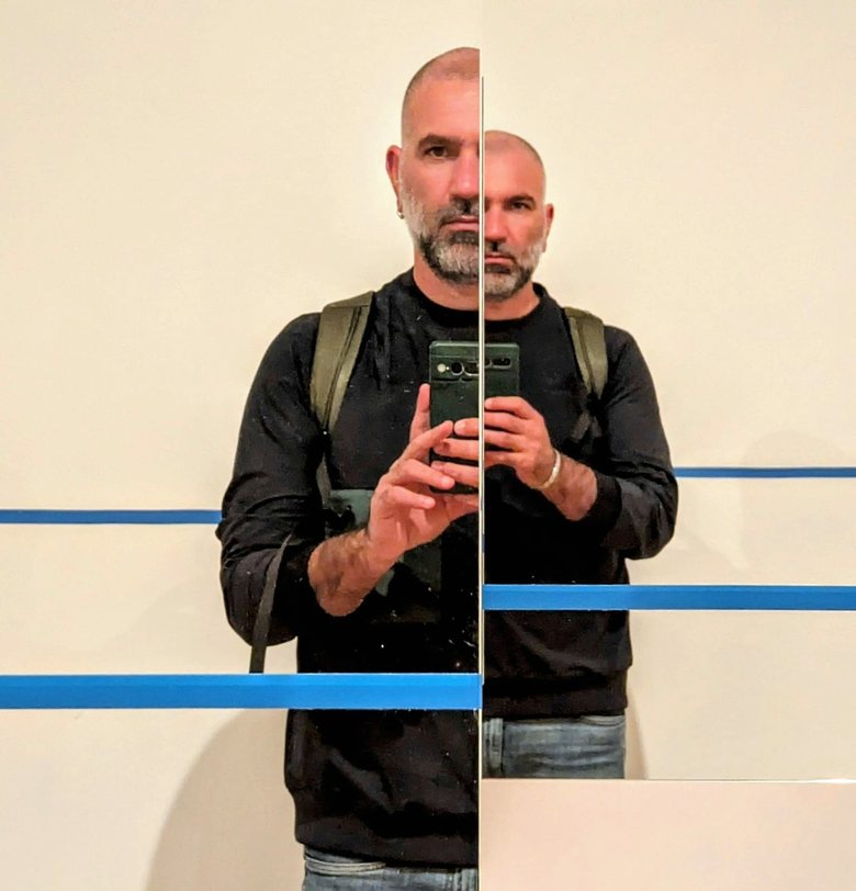 Rocco Pulizzi riflesso in due specchi affiancati, che mostrano due metà sfalsate del suo volto.